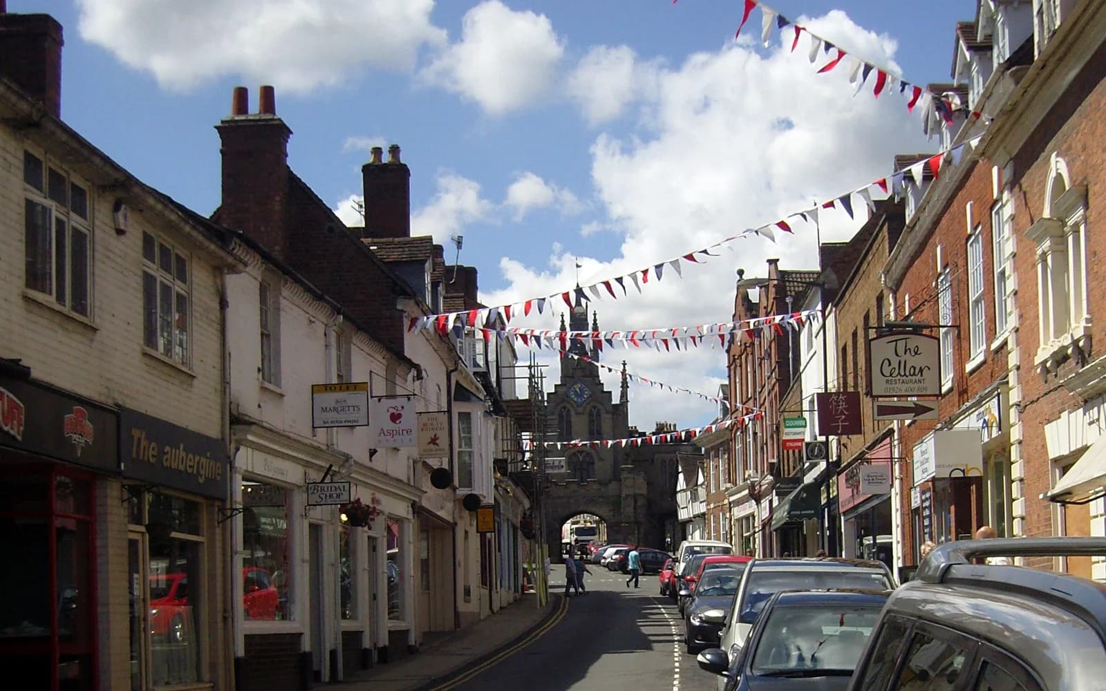 Smith Street in Warwick with historic shopfronts, parked cars, bunting, and a church tower under a bright sky.